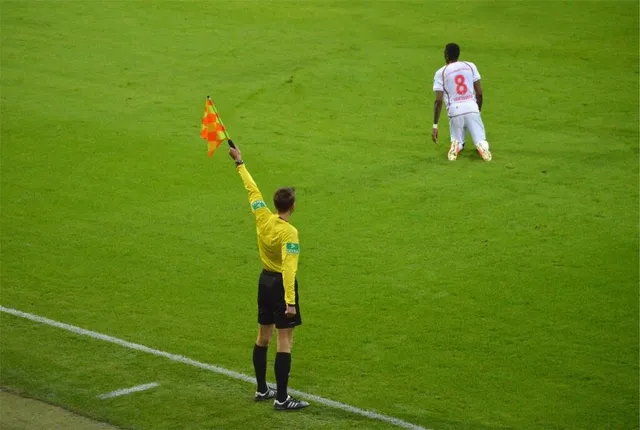 Un arbitru în echipament galben și negru ridică un steag de linie pe un teren de fotbal verde.