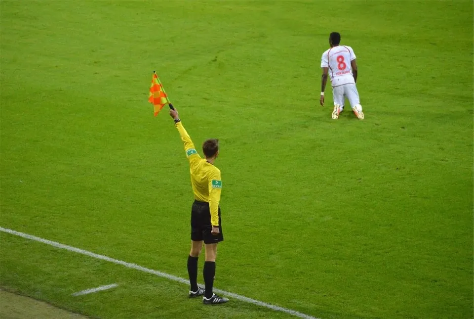 Un arbitru în echipament galben și negru ridică un steag de linie pe un teren de fotbal verde.