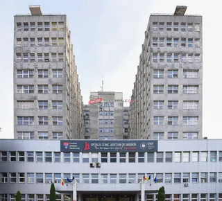 Două clădiri înalte de beton cu ferestre multiple încadrează un al treilea bloc de clădiri în fundal sub un cer înnorat.