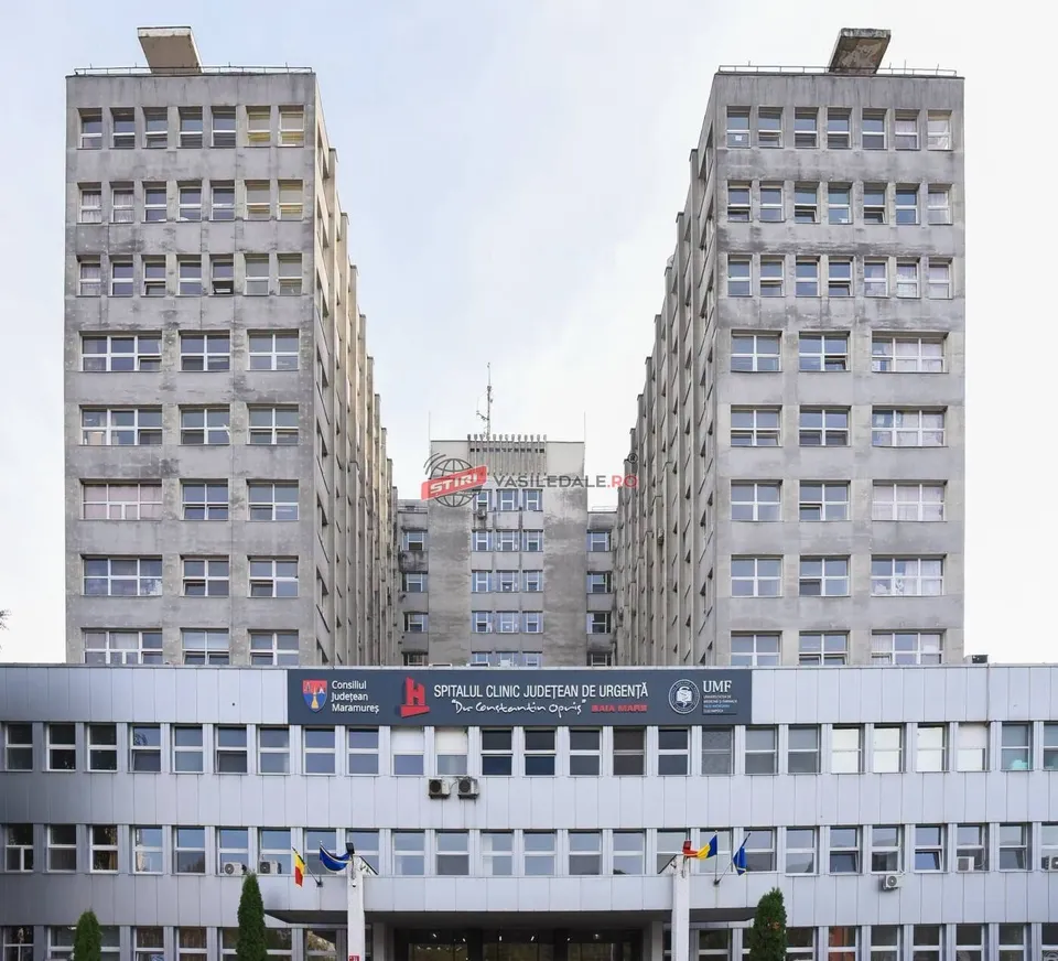 Două clădiri înalte de beton cu ferestre multiple încadrează un al treilea bloc de clădiri în fundal sub un cer înnorat.