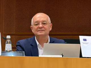Un bărbat cu ochelari și sacou albastru stă la o masă lângă un laptop și o sticlă de apă.