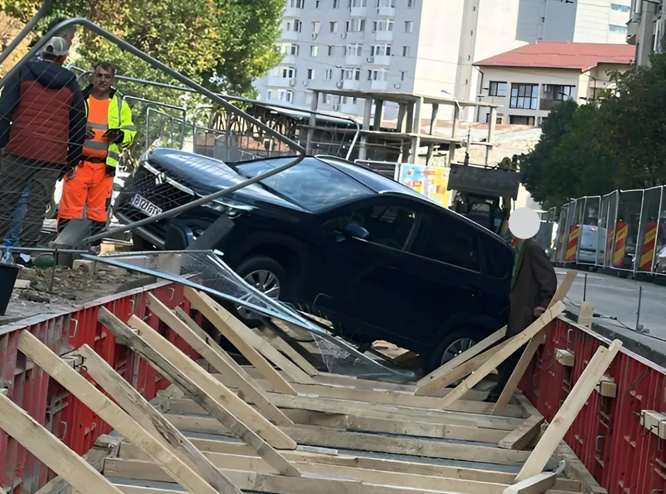 Un autovehicul negru este înclinat într-un șantier delimitat de bariere metalice și grinzi de lemn, în prezența unor persoane.