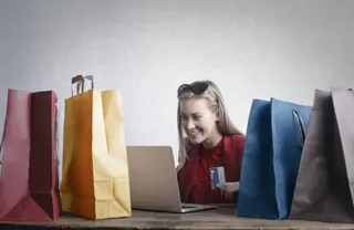 O femeie zâmbind stă la un birou cu un laptop deschis și mai multe sacoșe de cumpărături colorate în jurul ei.