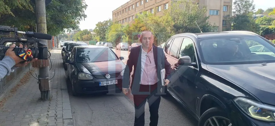 Un bărbat cu o cămașă roz și un sacou bordo merge pe lângă mașini parcate pe marginea unui drum.