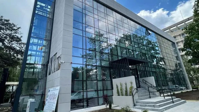 O clădire modernă cu fațadă din sticlă reflectă copacii și cerul deasupra unor scări de acces cu balustrade negre.