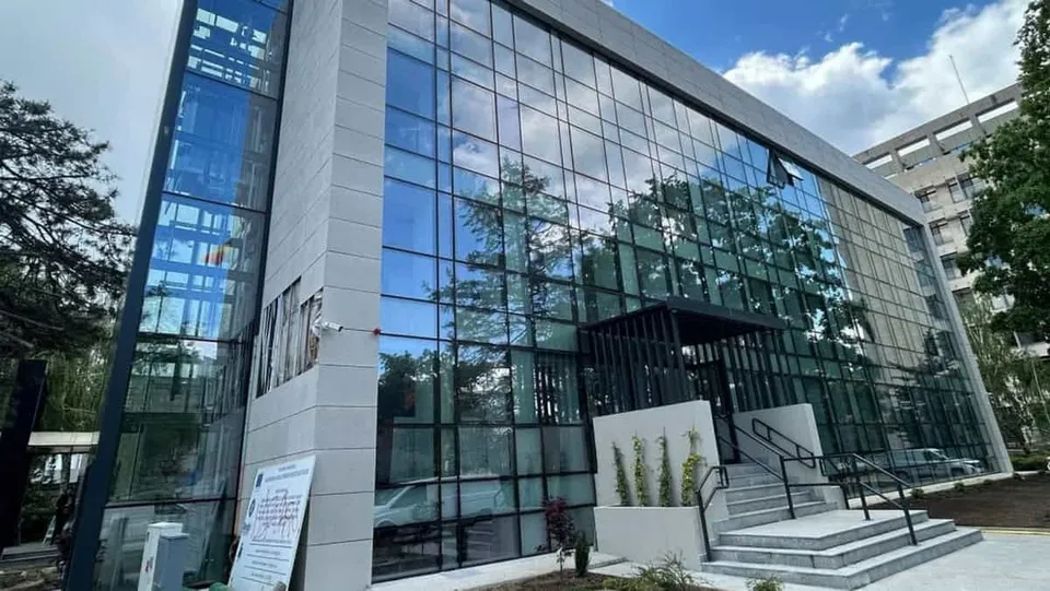 O clădire modernă cu fațadă din sticlă reflectă copacii și cerul deasupra unor scări de acces cu balustrade negre.