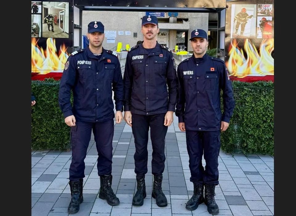 Trei bărbați îmbrăcați în uniforme de serviciu albastre și căciuli de aceeași culoare stau în picioare pe un trotuar lângă un gard de vegetație.