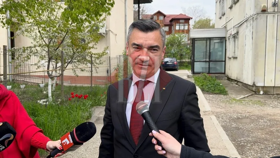 Un bărbat într-un costum negru și cămașă albă cu cravată roșie stă în fața unor microfoane pe un drum pietonal lângă clădiri și copaci.