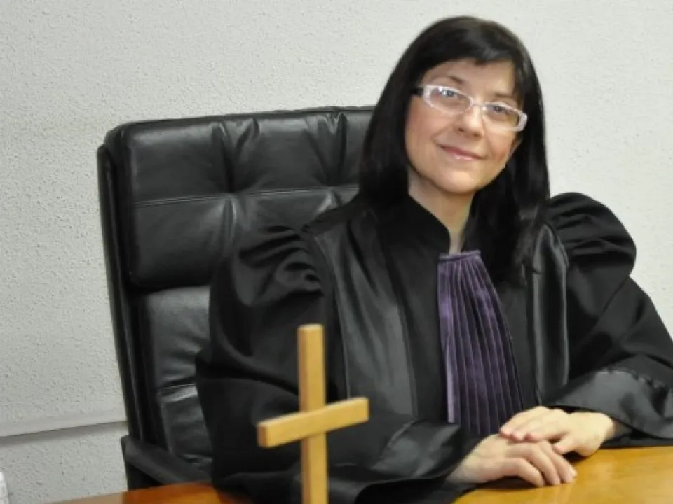 O femeie cu ochelari și păr negru poartă o robă neagră cu un element violet, stând la o masă de lemn în fața unui scaun negru.
