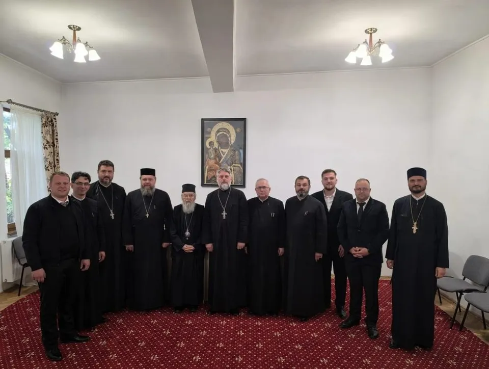 Un grup de unsprezece bărbați stau în rând într-o încăpere cu covor roșu și o icoană pe perete.