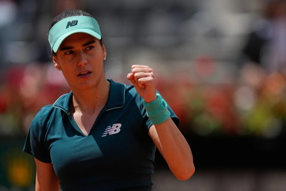 O femeie cu vizieră și tricou închis la culoare stă pe un teren de tenis cu pumnul strâns lângă față.