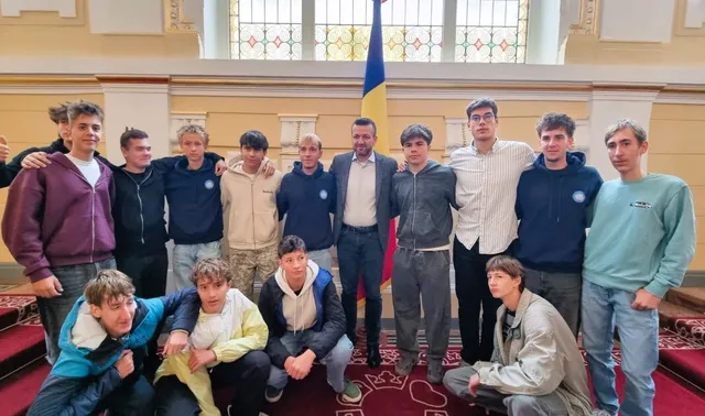 Un grup de tineri stă poziționat în două rânduri în fața unei ferestre mari, într-o încăpere cu pereți galbeni și un steag.
