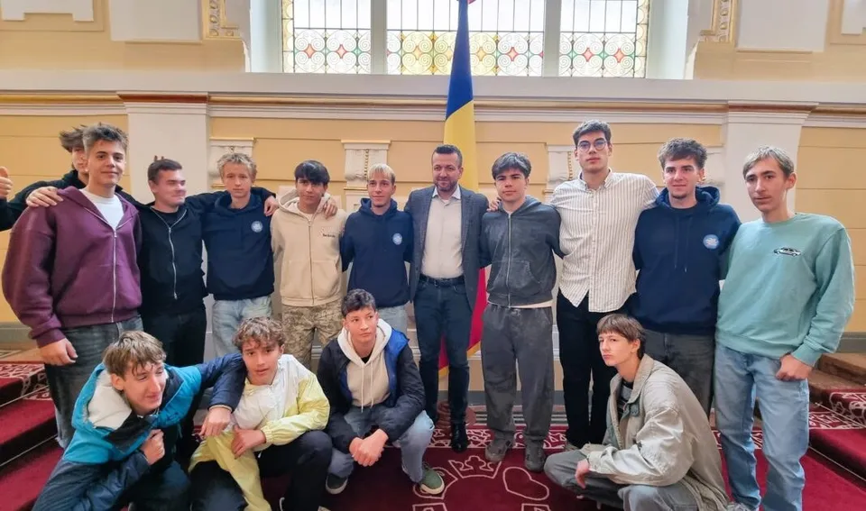 Un grup de tineri stă poziționat în două rânduri în fața unei ferestre mari, într-o încăpere cu pereți galbeni și un steag.