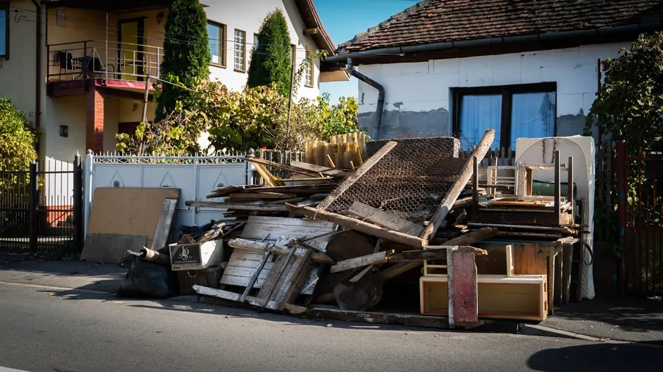 Un morman de obiecte din lemn, plăci și resturi de mobilier este depus pe marginea unui drum lângă o clădire albă.