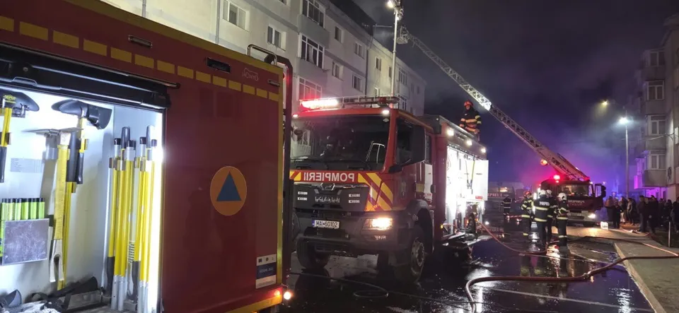 Un camion de pompieri roșu este parcat pe un drum umed, lângă un vehicul de intervenție cu o scară extinsă spre un bloc de locuințe.