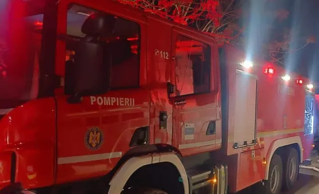 Un camion de pompieri roșu cu inscripția POMPIERII este parcat pe un drum noaptea sub lumini puternice.