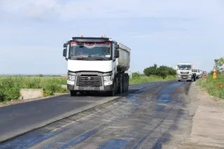 Un camion alb cu remorcă de tip basculant circulă pe un drum asfaltat lângă vegetație sub un cer înnorat.
