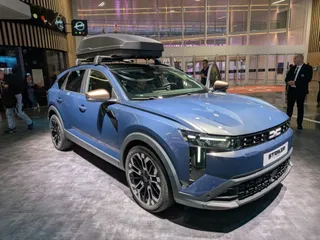 Un crossover albastru cu accesorii de acoperiș este expus într-un spațiu interior cu podea gri și panouri de sticlă în fundal.