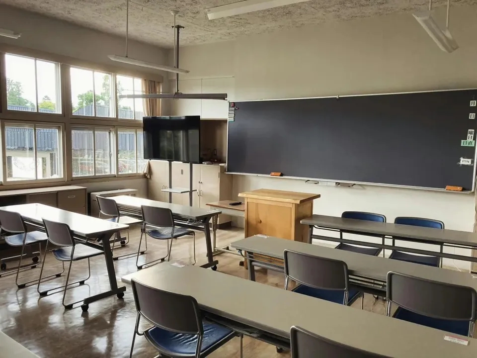O sală de clasă cu mai multe birouri gri și scaune negre dispuse în rânduri în fața unui panou negru mare pe perete.