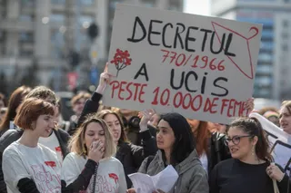 Un grup de femei strânge într-un spațiu public, în timp ce o persoană ridică un panou alb pe care scrie text cu marcaj negru și roșu.