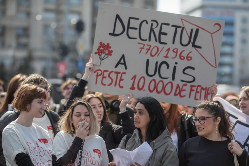 Un grup de femei strânge într-un spațiu public, în timp ce o persoană ridică un panou alb pe care scrie text cu marcaj negru și roșu.