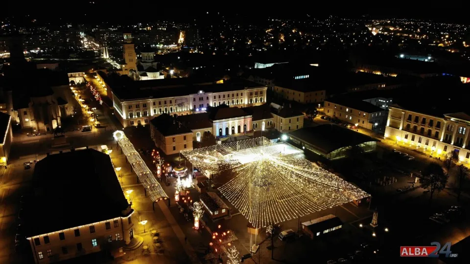 O vedere aeriană nocturnă a unei zone urbane cu clădiri iluminate și o structură decorativă luminoasă de formă triunghiulară în centrul unei piețe.