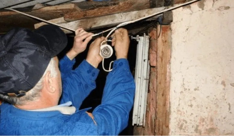 Un bărbat cu șapcă și cămașă albastră manipulează cabluri electrice și un dispozitiv rotund lângă un profil metalic pe un perete de cărămidă.