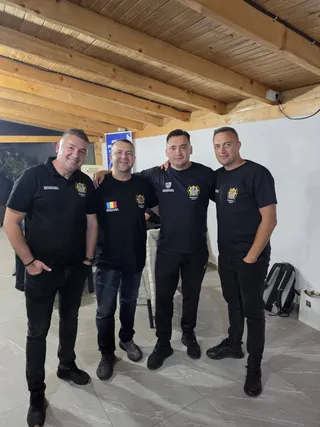 Patru bărbați în uniformă neagră cu logo-uri, în picioare și îmbrățișați, într-o cameră cu un acoperiș de lemn.