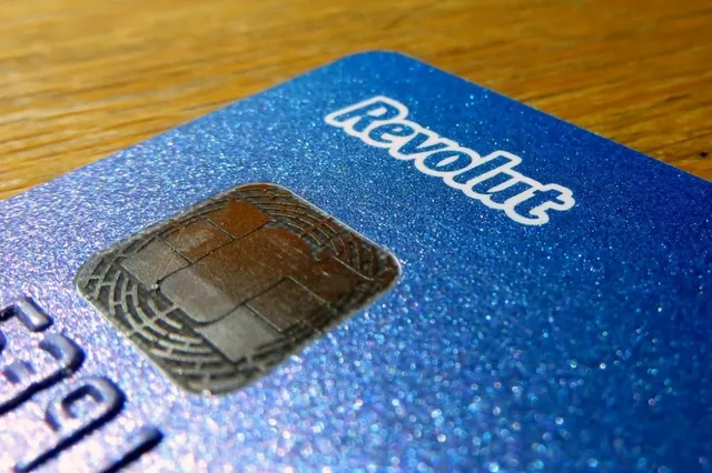 O parte dintr-o card bancar albastru cu textul Revolut și un cip metalic este vizibilă pe o suprafață de lemn.