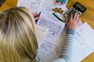 O persoană cu părul blond stă la o masă de lemn și folosește un calculator lângă diverse documente și monede.
