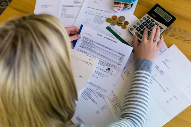 O persoană cu părul blond stă la o masă de lemn și folosește un calculator lângă diverse documente și monede.