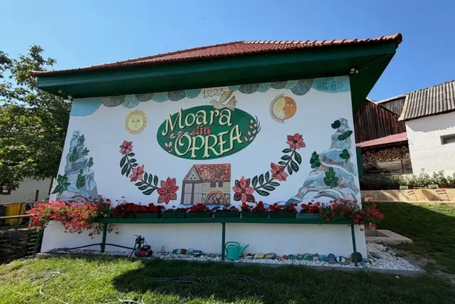 Un perete alb de o clădire prezintă un mural colorat cu flori, plante, un soare, o lună și textul Moara din Oprea.