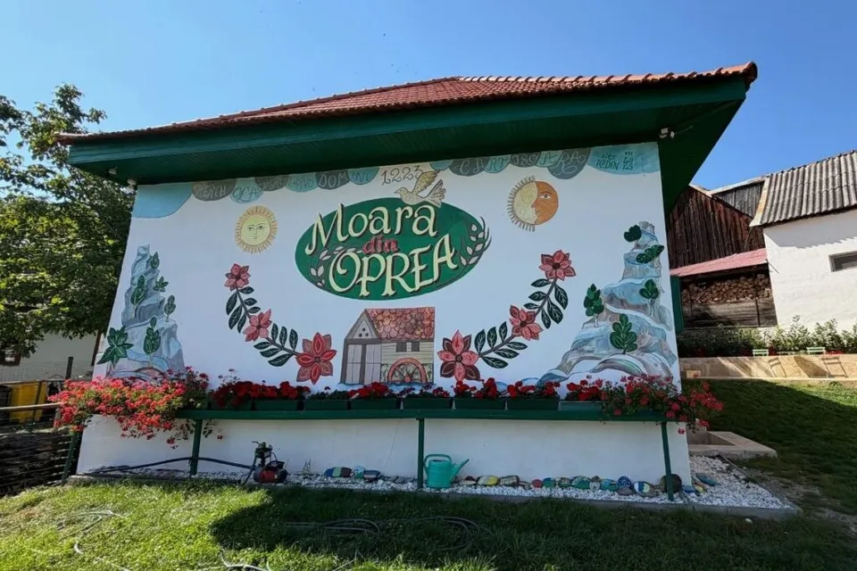 Un perete alb de o clădire prezintă un mural colorat cu flori, plante, un soare, o lună și textul Moara din Oprea.