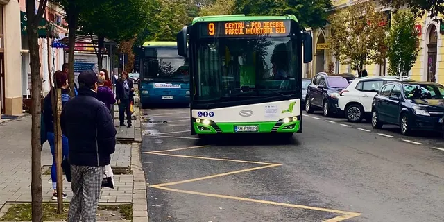 Un autobuz verde și alb cu numărul 9 circulă pe un drum asfaltat lângă mașini parcate și câțiva oameni pe trotuar.