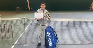 Un tânăr tenismen în vârstă de 12 ani, ținând un trofeu și un certificat, în picătură lângă o rețea de tenis, cu un ghiveci Yonex alăturat.