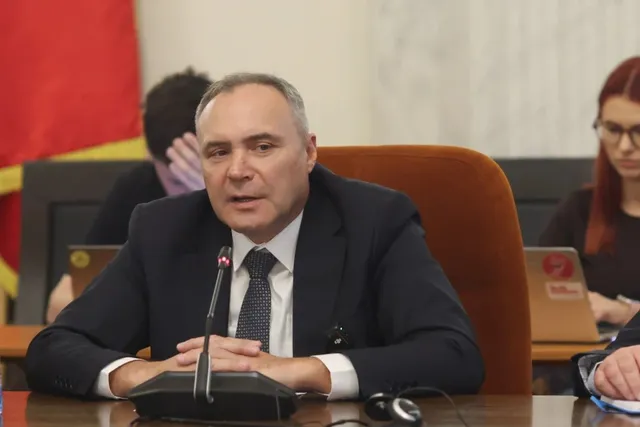 Un bărbat cu părul scurt și costum închis de culoare stă la o masă cu un microfon în față.