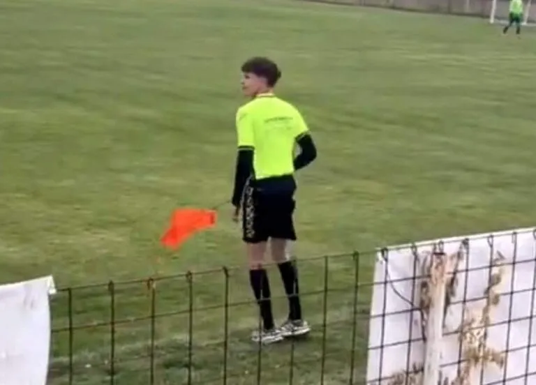 Un bărbat cu tricou galben și pantaloni scurți negri stă pe marginea unui teren de fotbal ținând un fanion portocaliu.