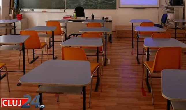 O sală de clasă cu mai multe birouri gri și scaune portocalii dispuse pe un podea de lemn.