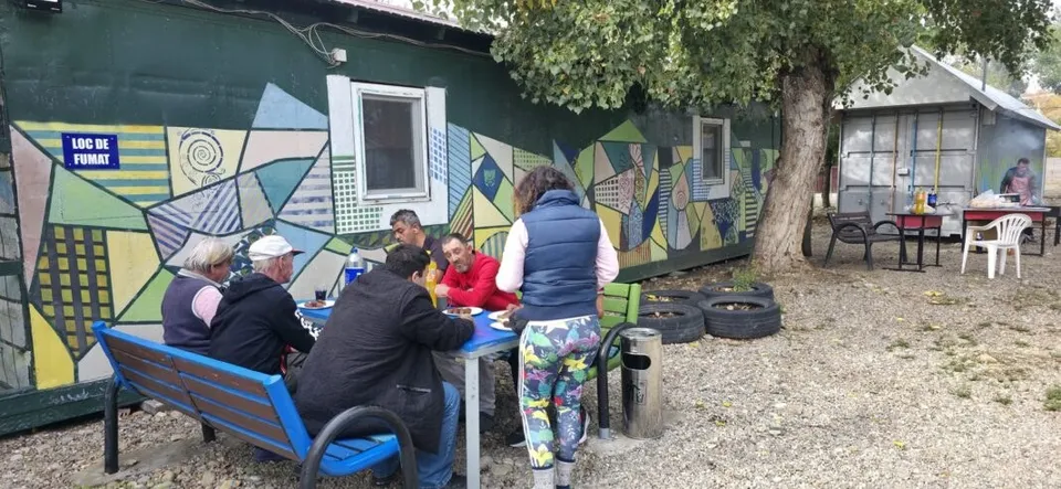 Oamenii stau la o masă de exterior lângă un container decorat cu grafitti colorate, în timp ce o persoană stă în picioare lângă ei.