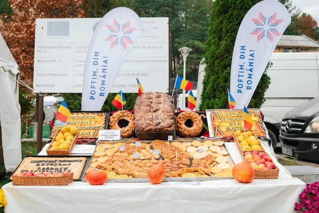 Diverse produse agricole și produse de panificație sunt expuse pe o masă acoperită cu un material alb, în fața unor steaguri și panouri informative.
