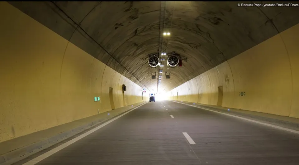 O autostradă cu două benzi traversează un tunel cu pereți galbeni și un plafon curbat dotat cu ventilatoare și lumini.