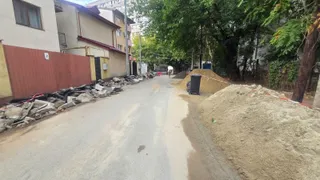 O stradă asfaltată este delimitată de grămezi de nisip, pietriș și resturi de beton pe margini, sub copaci verzi și clădiri rezidențiale.