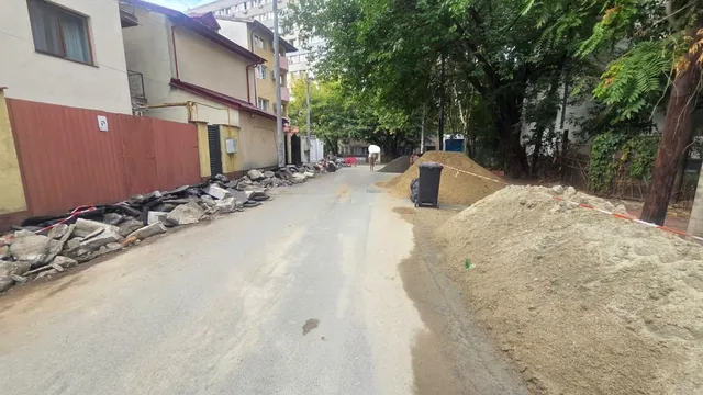 O stradă asfaltată este delimitată de grămezi de nisip, pietriș și resturi de beton pe margini, sub copaci verzi și clădiri rezidențiale.