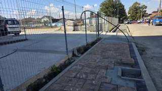 Un trotuar cu plăci de pavaj parțial dezasamblate se află lângă un gard de sârmă și o stradă asfaltată sub un cer senin.
