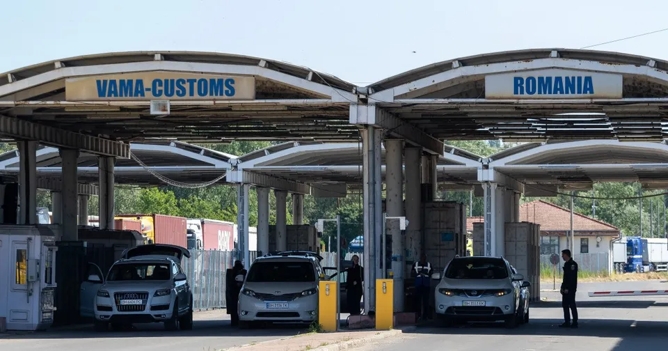 Vehiculele trec printr-un punct de frontieră cu două structuri acoperite, având semnele VAMA-CUSTOMS și ROMANIA deasupra.