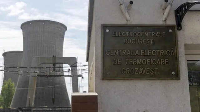 O placă metalică cu text este montată pe un perete gri, lângă două turnuri de răcire înalte sub un cer înnorat.