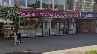 O clădire cu vitrine mari de sticlă prezintă un panou roșu pe care scrie Galeriile de Artă Lascăr Vorel, în fața căreia merg două persoane.