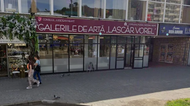 O clădire cu vitrine mari de sticlă prezintă un panou roșu pe care scrie Galeriile de Artă Lascăr Vorel, în fața căreia merg două persoane.
