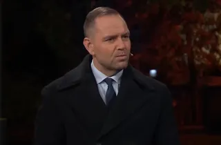 Un bărbat cu părul scurt, purtând haine negre, cămașă albastră și cravată, privește spre lateral într-un cadru cu fundal întunecat.
