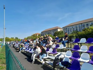 Oamenii stau așezați pe scaune de culoare albastră și albă în tribunele unui stadion sub un cer senin.